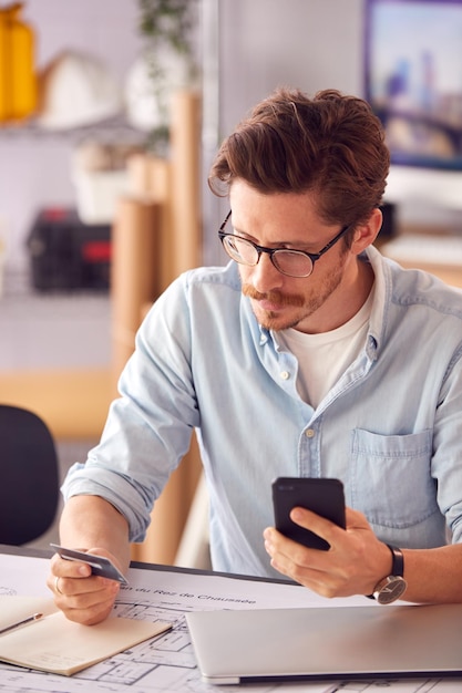 Mannelijke architect in kantoor werken aan bureau online aankopen doen met creditcard op mobiele telefoon