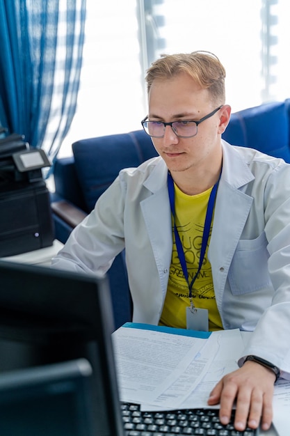 Mannelijke arts die aan medische expertise werkt terwijl hij aan het bureau achter de computer zit. Jonge dokter op kantoor.
