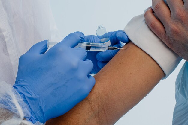 Foto mannelijke arts die het covid-19-vaccin in de spuit injecteert bij de patiënt van dichtbij. man met witte jas en latex handschoenen. concept van de bestrijding van het coronavirus. grijze achtergrond met turkoois licht. studio shoot