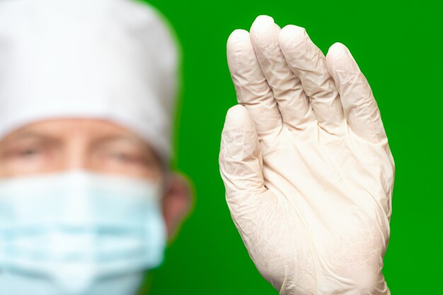 Foto mannelijke arts in chirurgie gezichtsmasker op defocus voorgrond zet palm van zijn hand naar voren in beschermende medische handschoen voorgrond, zwaait met zijn hand, stop of hallo teken. concept van bescherming op groene achtergrond.