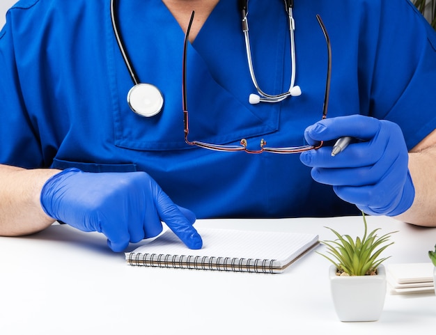Mannelijke arts in een blauw uniform zit aan een witte tafel en schrijft in een papieren notitieblok, steriele handschoenen aan zijn handen dragen, concept van het ontvangen van patiënten op kantoor