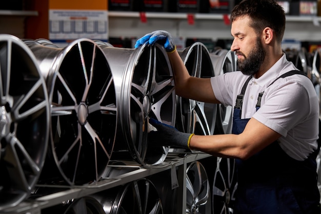 Mannelijke automonteur controleren nieuwe banden en schijven in autowinkel, voorbereiding voor verkoop, auto-industrie, bebaarde man in uniform op de werkplek