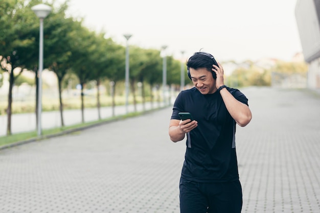 Mannelijke Aziatische atleet op een ochtendrun in het park bij het stadion luistert naar muziek en podcast in grote koptelefoons gebruikt de telefoon
