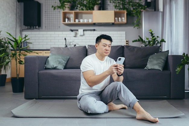Mannelijke Aziatische atleet thuis met behulp van telefoon zittend op de vloer tijdens ochtendoefening atleet in woonkamer
