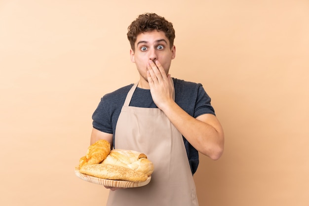 Mannelijke bakker die een lijst met verscheidene broden houdt die op beige muur met verrassingsgelaatsuitdrukking wordt geïsoleerd