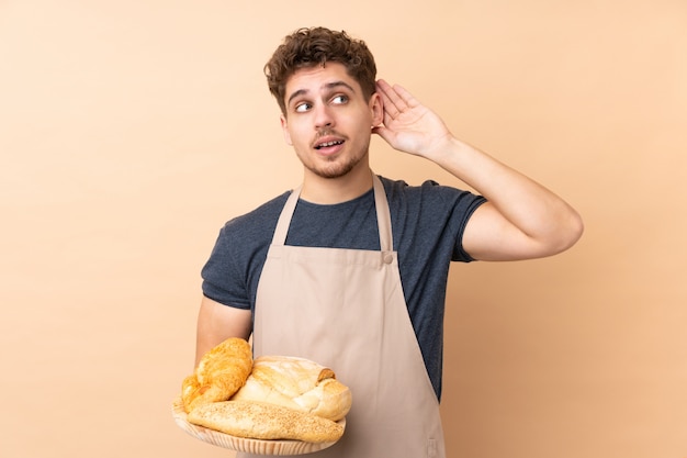 Mannelijke bakker die een lijst met verscheidene die brood houden op beige muur wordt geïsoleerd luisterend iets