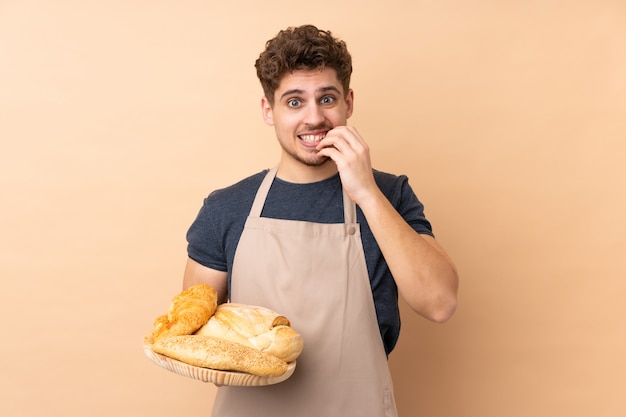 Mannelijke bakker die een lijst met verscheidene die brood houden op beige zenuwachtig en doen schrikken muur wordt geïsoleerd