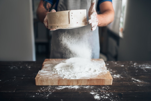 Mannelijke bakkershanden filteren de bloem door een zeef over een snijplank. Brood voorbereiding. Zelfgemaakte bakkerij