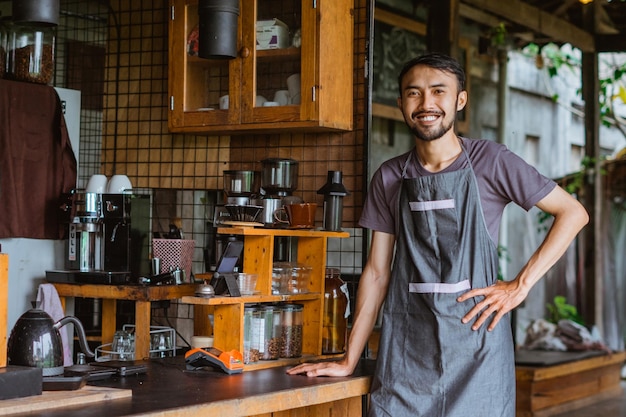 Mannelijke barista in schort staand met een glimlach en leunend op het barbureau met zijn hand op zijn middel