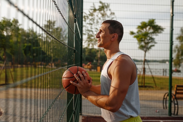 Mannelijke basketbalspeler met bal die zich bij het gaasomheining op openluchthof bevindt