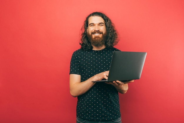 Mannelijke bebaarde hipster houdt een laptop vast en glimlacht naar de camera in een studio