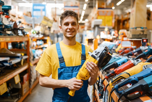 Mannelijke bouwer die haakse slijper kiest op de plank in de ijzerhandel. Constructor in uniforme blik op de goederen in de doe-het-zelfwinkel