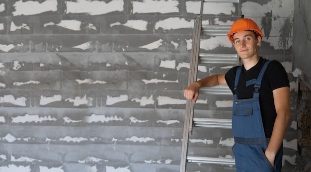Mannelijke bouwer gekleed in werkkleding en een oranje helm. staat bij de muur, leunend tegen de trap. Ruimte kopiëren