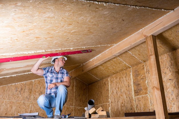 Mannelijke bouwvakker bouwer gehurkt op verhoogde steiger met behulp van rood niveau om de hoogte van het plafond in onvoltooide huis met blootgestelde multiplex spaanplaat te meten