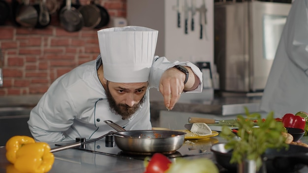 Mannelijke chef-kok giet geraspte kaas op gekookt voedsel in koekenpan, met behulp van rasp en keukengerei om gastronomische schotel te koken. Professionele kok in wit uniform die geraspte Parmezaanse kaas toevoegt aan culinaire maaltijd.