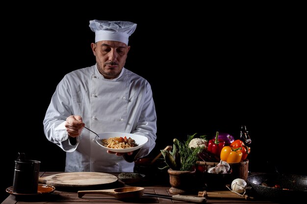 Mannelijke chef-kok in wit uniform bereidt spaghetti met groenten op het gerecht voordat hij serveert terwijl hij in de keuken van een restaurant werkt