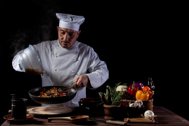 Mannelijke chef-kok in wit uniform giet gekookte spaghetti in pan wok voor het koken van pasta met groenten. Backstage van het bereiden van traditionele Italiaanse schotel op zwarte achtergrond