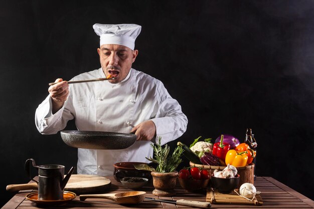 Mannelijke chef-kok in wit uniform proeft voedsel met behulp van een houten pollepel in de keuken van een restaurant