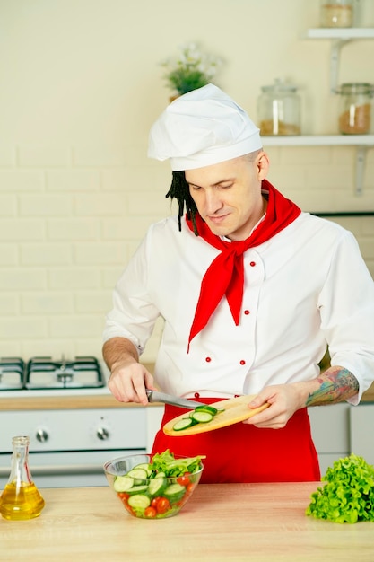 Mannelijke chef-kok met dreadlocks die verse plakjes komkommer toevoegen in glazen kom met salade in de keuken Een man kookt Vitaminen dieet- en sportvoeding Vegetarisch eten