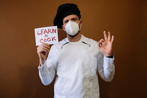 Mannelijke chef-kok met gezichtsmasker en een bord dat zegt: leren en koken