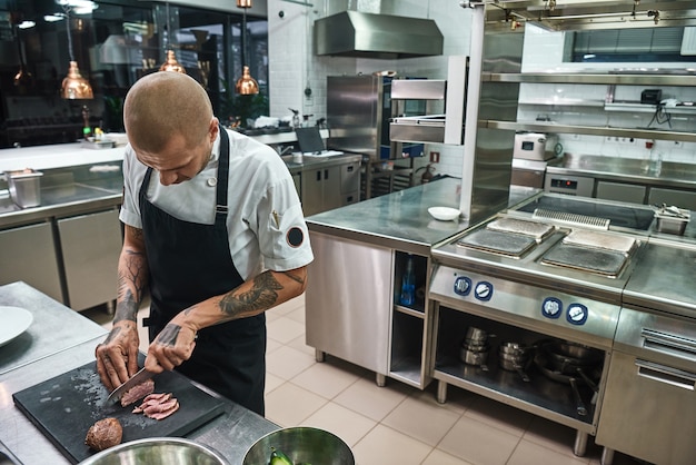 mannelijke chef-kok met prachtige tatoeages op zijn handen die vlees snijden terwijl hij in de keuken van een restaurant staat