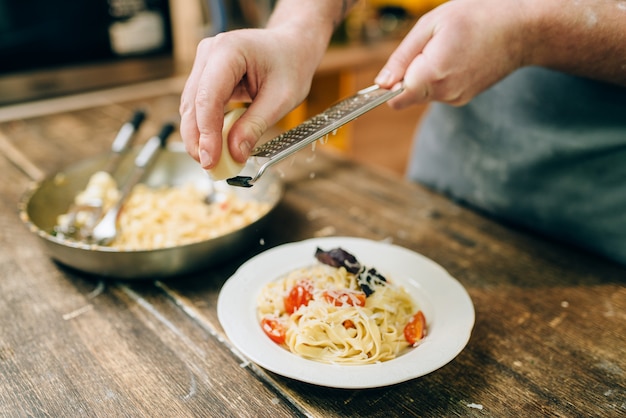 Mannelijke chef-kok raspt kaas in de plaat met vers gekookte fettuccine, pan op houten keukentafel. zelfgemaakte pasta bereidingsproces. traditionele italiaanse keuken