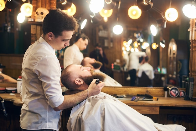 Mannelijke cliënt met baard zittend in kappersstoel Ernstige man met lange bruine baard Moderne populaire houthakkersstijl