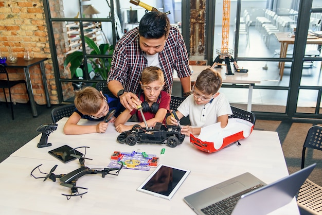 Mannelijke elektronische ingenieur met Europese schoolkinderen die in slim schoollaboratorium werken en model van radiografisch bestuurbare elektrische auto testen.