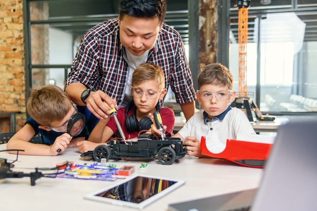 Mannelijke elektronische ingenieur met Europese schoolkinderen die in slim schoollaboratorium werken en model van radiografisch bestuurbare elektrische auto testen.