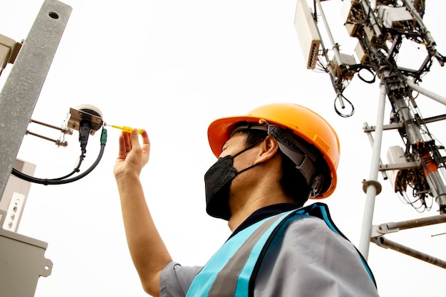 Mannelijke elektrotechnisch ingenieur die een helm draagt, gebruikt een schroevendraaier om de sensoren van de telecommunicatietoren te inspecteren.