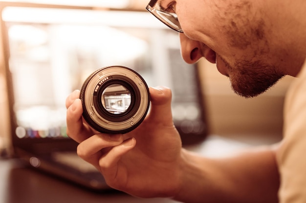 Foto mannelijke fotograaf heeft een lens in zijn hand en kijkt ernaar laptop op de onscherpe achtergrond