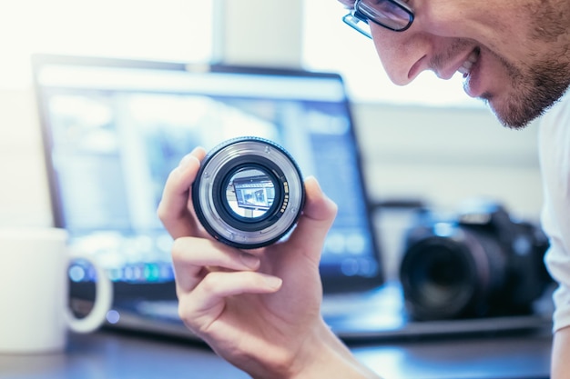 Mannelijke fotograaf heeft een lens in zijn hand en kijkt ernaar Laptop op de onscherpe achtergrond