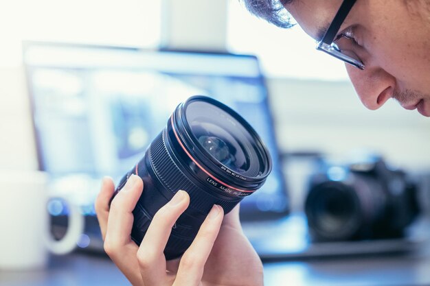 Mannelijke fotograaf heeft een lens in zijn hand en kijkt ernaar Laptop op de onscherpe achtergrond