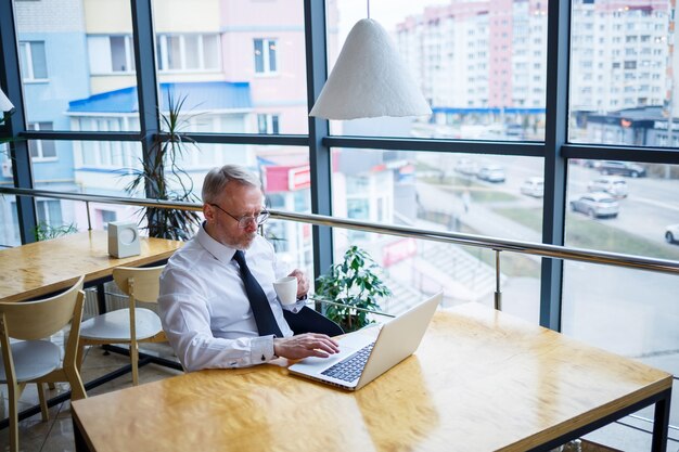 Mannelijke freelancer werkt in een café aan een nieuw zakelijk project. Zit aan een groot raam aan de tafel. Kijkt naar een laptopscherm met een kopje koffie