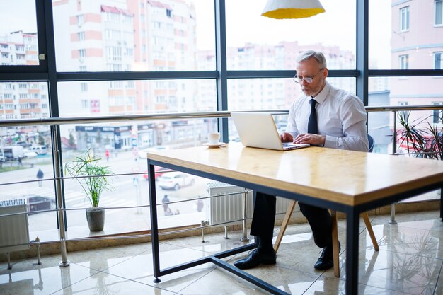 Mannelijke freelancer werkt in een café aan een nieuw zakelijk project. Zit aan een groot raam aan de tafel. Kijkt naar een laptopscherm met een kopje koffie