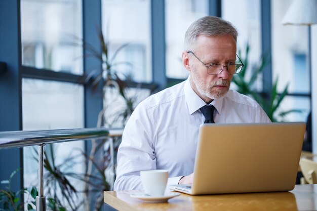 Mannelijke freelancer werkt in een café aan een nieuw zakelijk project. Zit aan een groot raam aan de tafel. Kijkt naar een laptopscherm met een kopje koffie