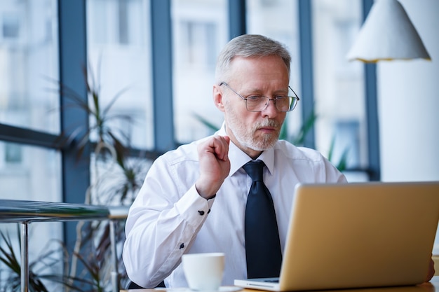 Mannelijke freelancer werkt in een café aan een nieuw zakelijk project. Zit aan een groot raam aan de tafel. Kijkt naar een laptopscherm met een kopje koffie