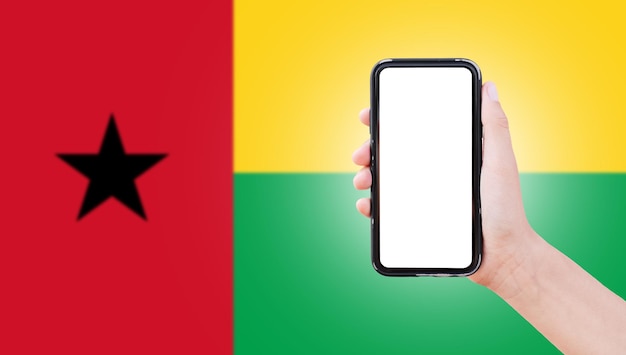 Mannelijke hand met smartphone met blanco scherm op de achtergrond van wazige vlag van Guinee-Bissau Close-upweergave