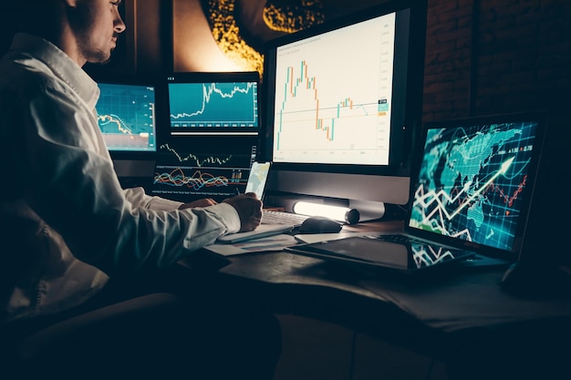 Foto mannelijke handelaar die beursgegevens analyseert terwijl hij op een kantoorwerkstation zit met meerdere computers