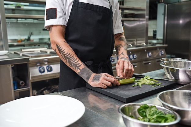 mannelijke handen met mooie tatoeages die komkommer pellen voor salade terwijl ze in de keuken van een restaurant staan