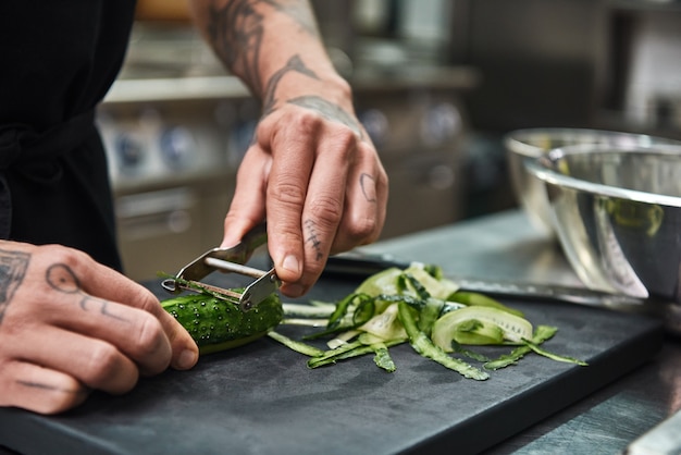 mannelijke handen met tatoeages die komkommer pellen voor salade terwijl ze in de keuken van het restaurant staan