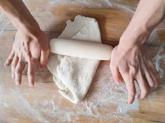 Mannelijke handen verspreiden deeg met een deegroller op houten keukentafel met bloem erop