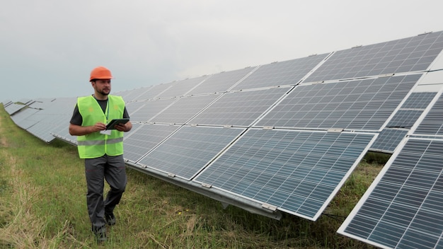 Mannelijke ingenieur in uniforme en harde helm loopt op hernieuwbare energiestation met digitale tablet en controleert de installatie van zonnepanelen. Alternatieve energie. Schone energieconcept.