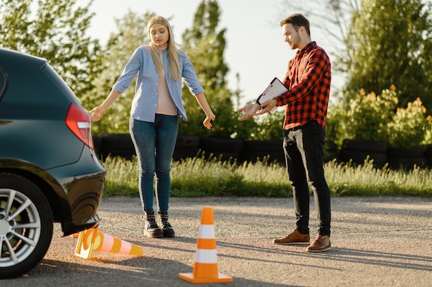 Mannelijke instructeur en vrouw bij de auto en neergehaalde kegel, les in rijschool. Man die dame leert voertuig te besturen. Rijbewijs opleiding