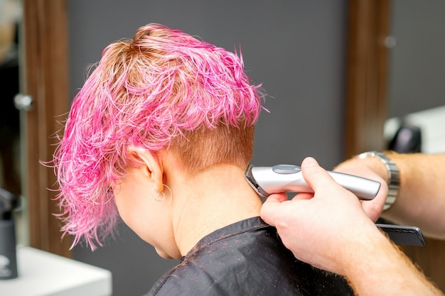 Mannelijke kapper scheert nek van een jonge blanke vrouw met een kort roze kapsel door elektrisch scheerapparaat in een kapsalon close-up