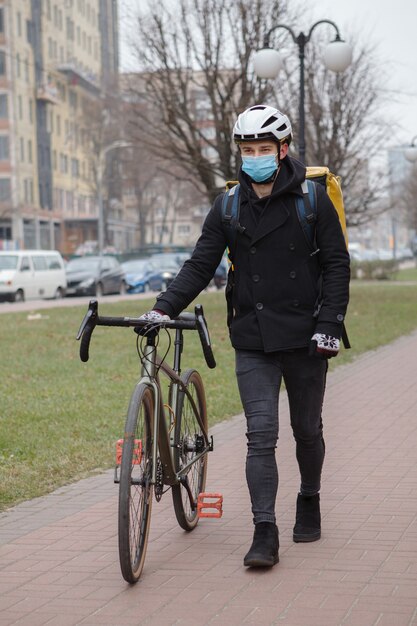 Mannelijke koerier die medisch gezichtsmasker draagt, met zijn fiets loopt, die thermo rugzak draagt