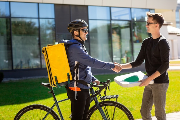 Mannelijke koerier met fiets en thermische tas geven een doos aan klant op straat in de buurt van het kantoor.