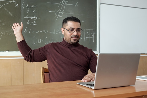 Mannelijke leraar van gemengd ras in vrijetijdskleding wijzend op de oplossing van de vergelijking op het bord terwijl hij aan het bureau zit en naar het laptopscherm kijkt