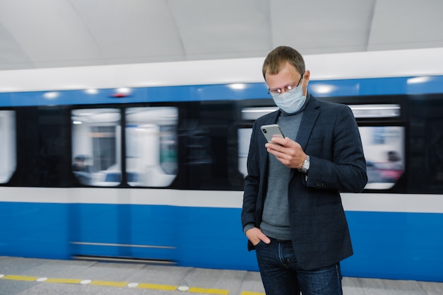 Mannelijke passagier draagt gezichtsmasker poseert op perron, wacht op trein, pendelt ondergronds, geconcentreerd in smartphoneapparaat, leest nieuws online. virusbewustzijn op openbare plaatsen. coronavirus-uitbraak