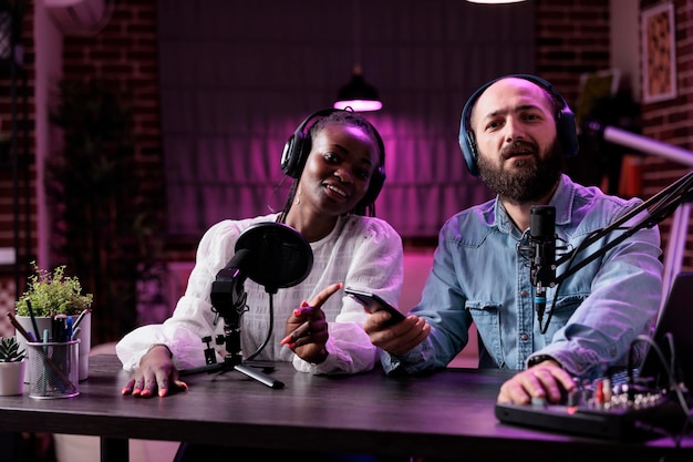Mannelijke presentator en gast opnemen podcast vlog-discussie in studio met neonlichten. Multi-etnisch team van mensen die videogesprekken filmen op camera, uitzenden op sociale media.